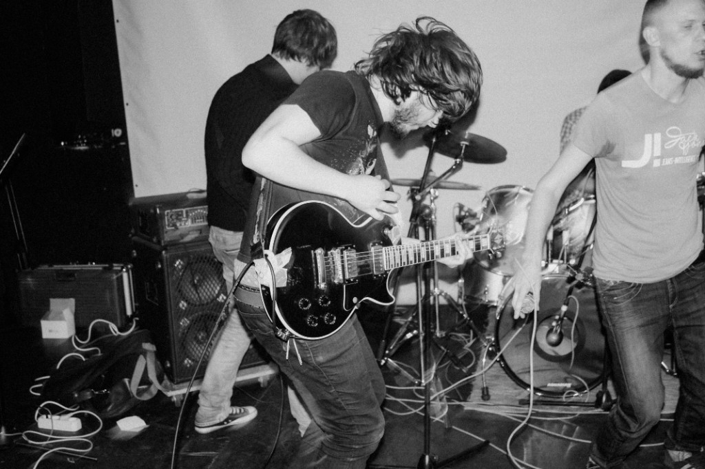 Band playing on a small club stage with guitar, drums, and amplifiers